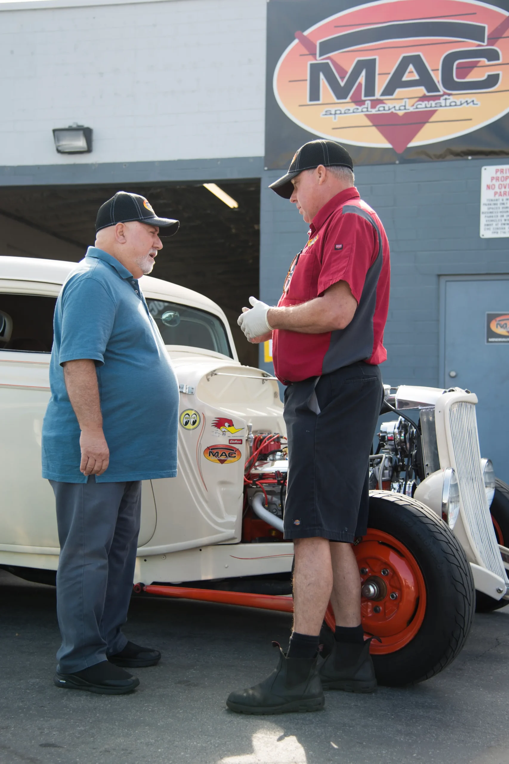 Two men in MAC caps talking in front of a white hot rod outside the MAC Speed and Custom shop